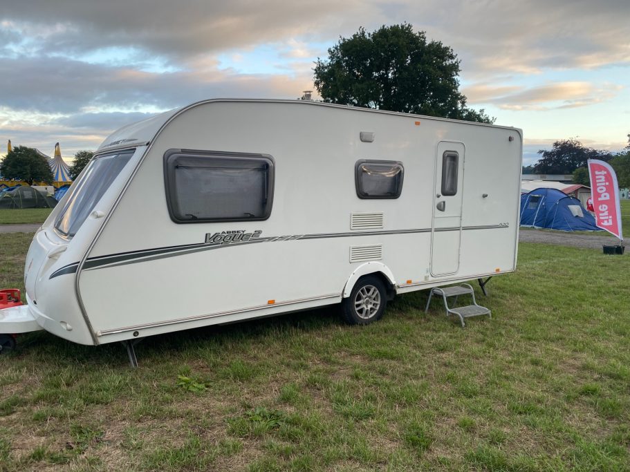 White caravan parked on grass, with a tent in the background and trees nearby.
