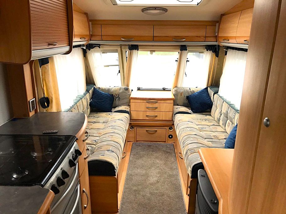 Interior of a caravan featuring two seating areas, cupboards, and a kitchen space.