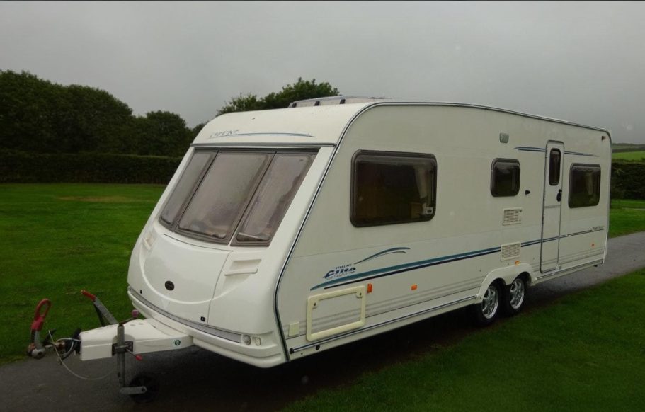 White caravan parked on a road, surrounded by greenery under a cloudy sky.